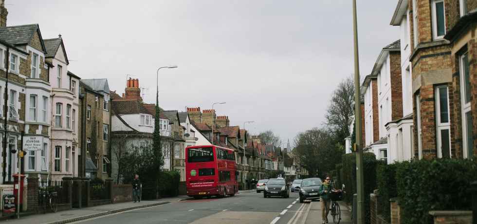 A red double decker bus on a street with cars and houses.